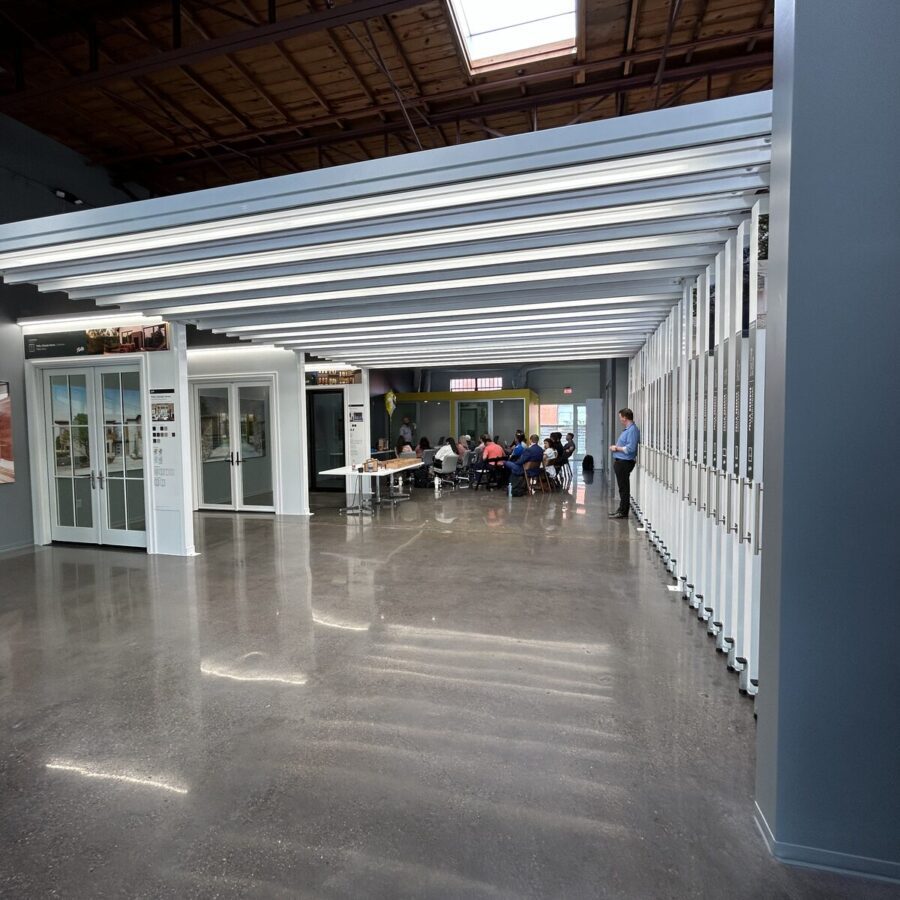 A group of people sit at tables in a meeting area inside a modern building with polished concrete floors and exposed ceiling beams.
